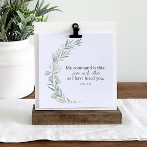 May include: A white framed sign with a wood base, featuring a floral design and the text "My command is this: Love each other as I have loved you. John 13:34". The sign is sitting on a white table runner with a bouquet of lavender in a white vase.