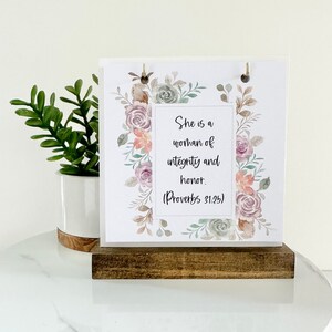 May include: A white card with a floral design and the text "She is a woman of integrity and honor. (Proverbs 31:25)" on a wooden stand.