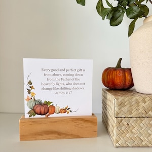 May include: A white card with the text "Every good and perfect gift is from above...James 1:17" sits in a wooden holder. The card features a watercolor illustration of pumpkins. A small orange pumpkin rests on a woven box.