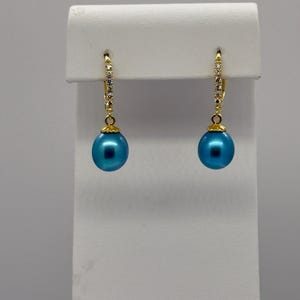May include: A pair of gold-colored drop earrings with a blue, spherical bead. The earrings have a small, jeweled accent on the top portion of the drop. The earrings are displayed on a white stand.