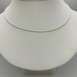 May include: A delicate silver necklace displayed on a white mannequin. The necklace features a fine chain with small, evenly spaced links. The jewelry is presented against a neutral background, highlighting its simple elegance.