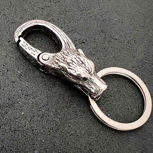 May include: A silver-colored keychain featuring a wolf head design. The keychain includes a carabiner-style clip and a split ring for keys. The wolf head is intricately detailed, and the metal has a polished finish.