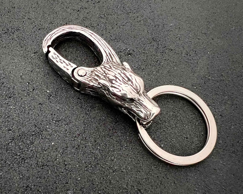 May include: A silver-colored keychain featuring a wolf head design. The keychain includes a carabiner-style clip and a split ring for keys. The wolf head is intricately detailed, and the metal has a polished finish.