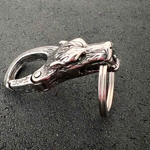 May include: A silver-colored metal carabiner clip with a wolf head design. The clip features a hinged gate and a key ring attachment. The wolf head is detailed with textured fur and facial features.