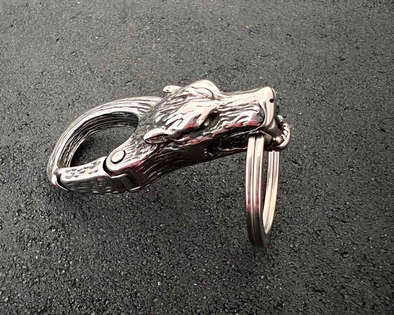 May include: A silver-colored metal carabiner clip with a wolf head design. The clip features a hinged gate and a key ring attachment. The wolf head is detailed with textured fur and facial features.