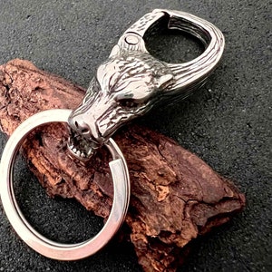 May include: A silver-colored keychain featuring a wolf head design. The wolf head is intricately detailed with visible teeth and fur texture. The keychain includes a large, round key ring and a spring-loaded clasp.