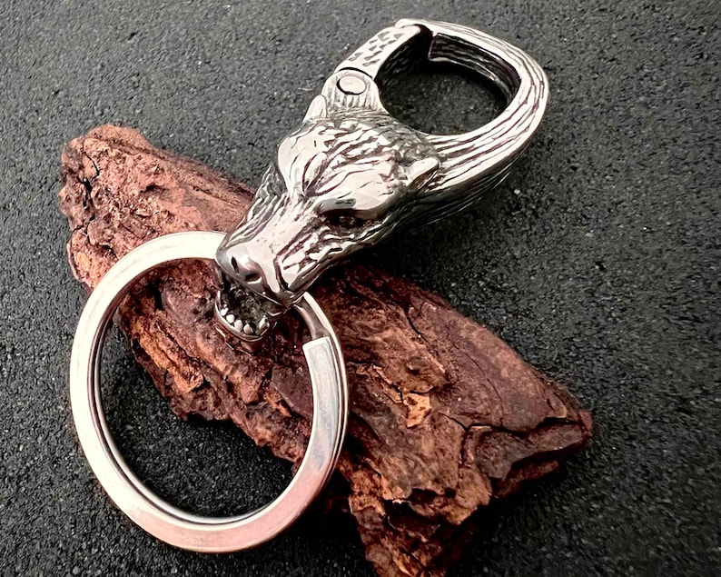 May include: A silver-colored keychain featuring a wolf head design. The wolf head is intricately detailed with visible teeth and fur texture. The keychain includes a large, round key ring and a spring-loaded clasp.