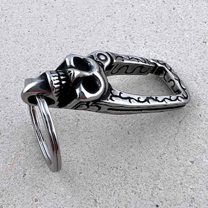 Puede incluir: Un llavero plateado con dise&ntilde;o de calavera. La calavera est&aacute; detallada con dientes y cuencas de los ojos visibles. El llavero tiene un asa decorativa estampada y un anillo met&aacute;lico para llaves.