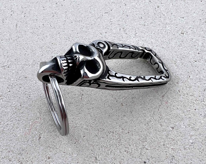 Puede incluir: Un llavero plateado con dise&ntilde;o de calavera. La calavera est&aacute; detallada con dientes y cuencas de los ojos visibles. El llavero tiene un asa decorativa estampada y un anillo met&aacute;lico para llaves.
