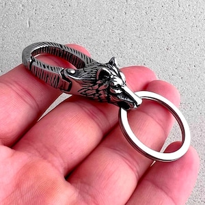 May include: A silver-colored keychain featuring a detailed wolf head design. The keychain has a sturdy clasp and a circular key ring. The wolf's head is intricately sculpted, showcasing fur and facial features. The metal has a polished finish.