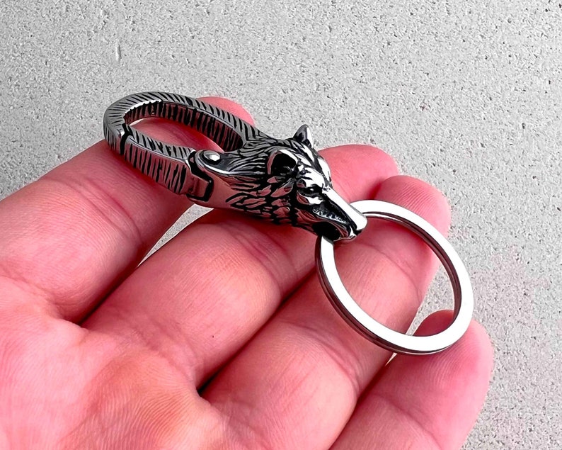 May include: A silver-colored keychain featuring a detailed wolf head design. The keychain has a sturdy clasp and a circular key ring. The wolf's head is intricately sculpted, showcasing fur and facial features. The metal has a polished finish.
