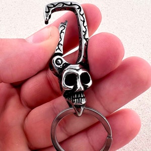 Puede incluir: Un llavero plateado con dise&ntilde;o de calavera. La calavera tiene cuencas oculares grandes y una mand&iacute;bula detallada. El llavero incluye un mosquet&oacute;n y una anilla para llaves.