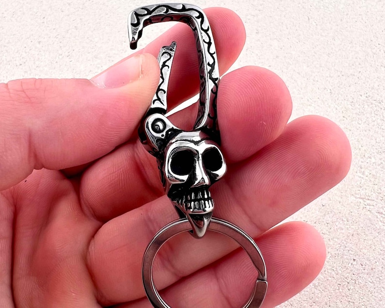 Puede incluir: Un llavero plateado con dise&ntilde;o de calavera. La calavera tiene cuencas oculares grandes y una mand&iacute;bula detallada. El llavero incluye un mosquet&oacute;n y una anilla para llaves.
