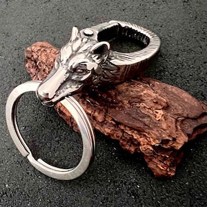 May include: A silver-colored keychain featuring a wolf's head design. The wolf's head is intricately detailed, with visible eyes and fur texture. The keychain includes a split ring for attaching keys and a spring-loaded clasp. The background is a dark, textured surface.