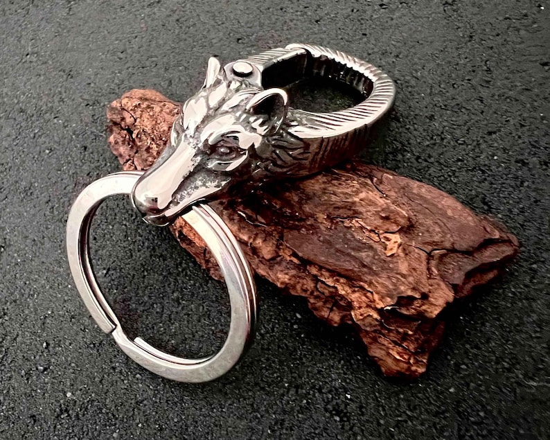 May include: A silver-colored keychain featuring a wolf's head design. The wolf's head is intricately detailed, with visible eyes and fur texture. The keychain includes a split ring for attaching keys and a spring-loaded clasp. The background is a dark, textured surface.