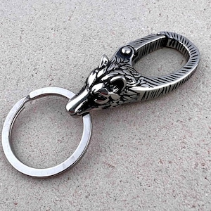 May include: A silver-colored keychain featuring a detailed wolf head design. The keychain includes a split ring and a spring-loaded clasp. The wolf head is intricately carved, showcasing the animal's features. The background is a textured, light gray surface.