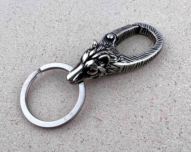 May include: A silver-colored keychain featuring a detailed wolf head design. The keychain includes a split ring and a spring-loaded clasp. The wolf head is intricately carved, showcasing the animal's features. The background is a textured, light gray surface.