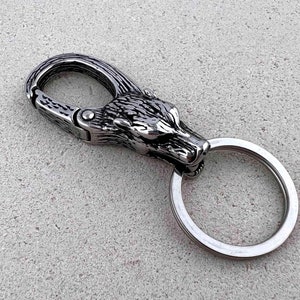 May include: A silver-colored keychain featuring a wolf head design. The keychain has a spring-loaded clasp and a split ring for keys. The wolf head is intricately detailed, with textured fur and sharp features. The metal has a polished finish.