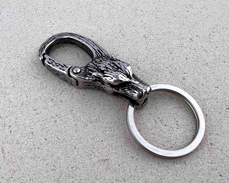 May include: A silver-colored keychain featuring a wolf head design. The keychain has a spring-loaded clasp and a split ring for keys. The wolf head is intricately detailed, with textured fur and sharp features. The metal has a polished finish.