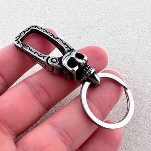 Puede incluir: Un llavero plateado con dise&ntilde;o de calavera. La calavera tiene dientes y cuencas de los ojos visibles. El llavero tiene un cierre con un patr&oacute;n ornamental y un anillo met&aacute;lico para llaves.