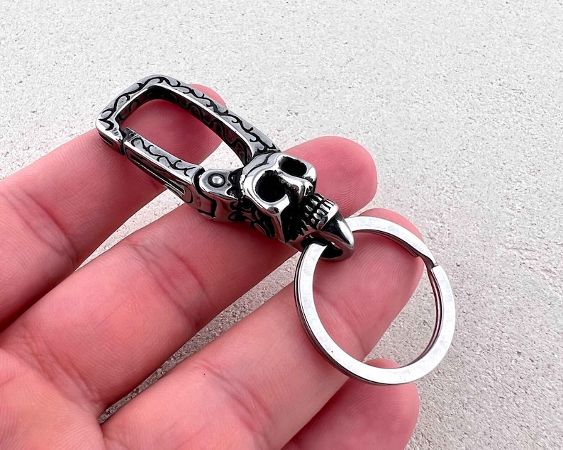 Puede incluir: Un llavero plateado con dise&ntilde;o de calavera. La calavera tiene dientes y cuencas de los ojos visibles. El llavero tiene un cierre con un patr&oacute;n ornamental y un anillo met&aacute;lico para llaves.