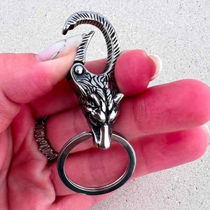 May include: A silver-colored keychain featuring a wolf head design. The keychain has a large, round ring and a spring-loaded clasp. The wolf head is intricately detailed with etched fur and facial features. The keychain is held in a hand.