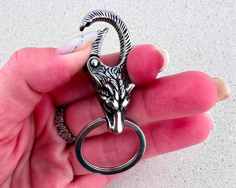 May include: A silver-colored keychain featuring a wolf head design. The keychain has a large, round ring and a spring-loaded clasp. The wolf head is intricately detailed with etched fur and facial features. The keychain is held in a hand.