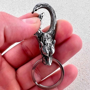 May include: A silver-colored keychain featuring a wolf head design. The keychain has a large, sturdy clasp and a circular key ring. The wolf head is intricately detailed, with textured fur and a fierce expression. The keychain is held in a hand.