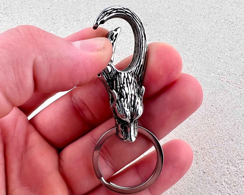 May include: A silver-colored keychain featuring a wolf head design. The keychain has a large, sturdy clasp and a circular key ring. The wolf head is intricately detailed, with textured fur and a fierce expression. The keychain is held in a hand.
