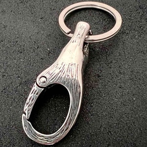 May include: A silver-colored keychain with a carabiner-style clasp and a key ring. The clasp has a textured surface and a hinged opening. The key ring is circular and made of the same shiny metal. The background is a dark, textured surface.