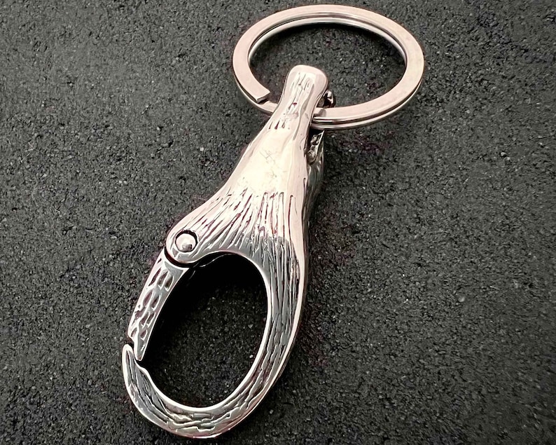 May include: A silver-colored keychain with a carabiner-style clasp and a key ring. The clasp has a textured surface and a hinged opening. The key ring is circular and made of the same shiny metal. The background is a dark, textured surface.