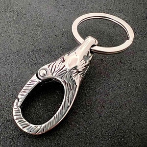 May include: A silver-colored keychain featuring a wolf head design. The keychain has a spring-loaded clasp and a split ring for keys. The wolf head is intricately detailed, with carved fur and facial features. The metal has a polished finish.