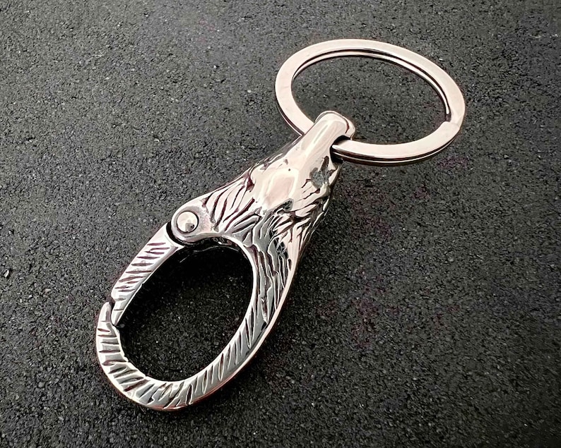 May include: A silver-colored keychain featuring a wolf head design. The keychain has a spring-loaded clasp and a split ring for keys. The wolf head is intricately detailed, with carved fur and facial features. The metal has a polished finish.