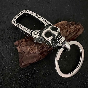Puede incluir: Un llavero plateado con forma de calavera con un dise&ntilde;o detallado. El llavero presenta una cabeza de calavera con cuencas de los ojos y dientes abiertos, sujeta a un cierre decorativo y un llavero. El cierre tiene un patr&oacute;n adornado.