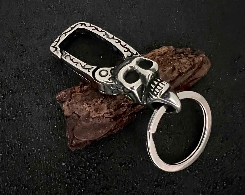 Puede incluir: Un llavero plateado con forma de calavera con un dise&ntilde;o detallado. El llavero presenta una cabeza de calavera con cuencas de los ojos y dientes abiertos, sujeta a un cierre decorativo y un llavero. El cierre tiene un patr&oacute;n adornado.