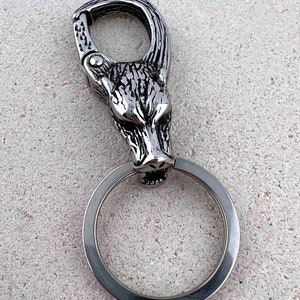 May include: A silver-colored keychain featuring a wolf head design. The wolf head is detailed with textured fur and a fierce expression. The keychain includes a large ring and a clasp for attaching to a belt loop or bag.