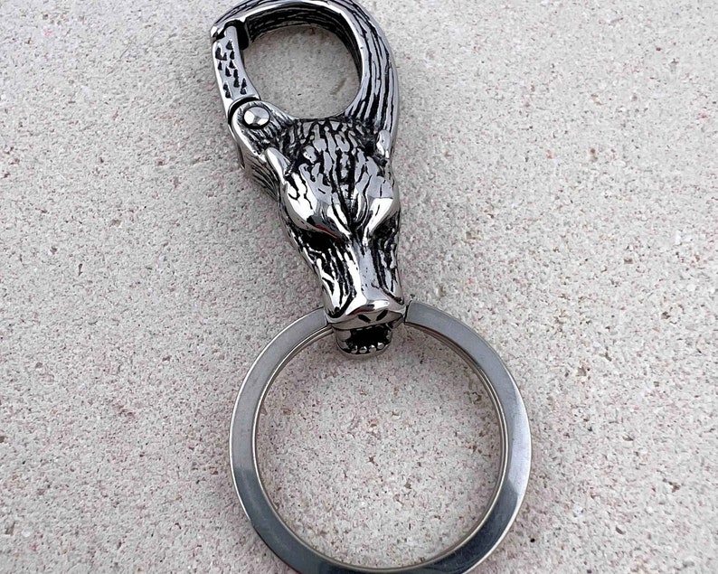 May include: A silver-colored keychain featuring a wolf head design. The wolf head is detailed with textured fur and a fierce expression. The keychain includes a large ring and a clasp for attaching to a belt loop or bag.
