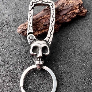 Puede incluir: Llavero plateado con dise&ntilde;o de calavera. La calavera presenta detalles faciales y un anillo para llaves. El mosquet&oacute;n tiene un patr&oacute;n ornamental. Llavero de metal.