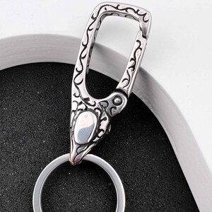 Puede incluir: Un llavero plateado tipo mosquet&oacute;n con un patr&oacute;n ornamental y ondulado. El llavero tiene un cierre de bisagra y un llavero circular. El dise&ntilde;o es detallado y parece estar hecho de metal.