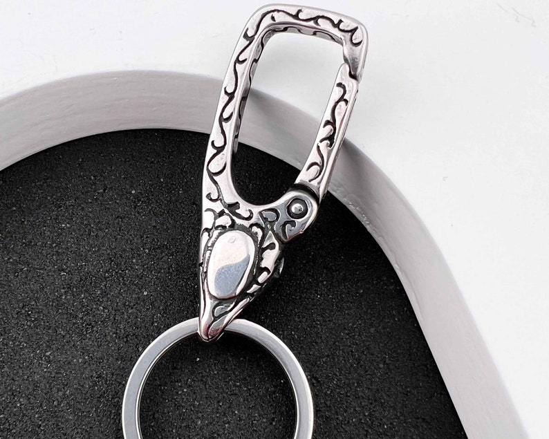 Puede incluir: Un llavero plateado tipo mosquet&oacute;n con un patr&oacute;n ornamental y ondulado. El llavero tiene un cierre de bisagra y un llavero circular. El dise&ntilde;o es detallado y parece estar hecho de metal.