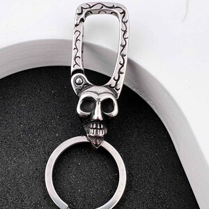 Puede incluir: Un llavero plateado con un colgante de calavera. La calavera tiene rasgos faciales detallados y est&aacute; unida a un llavero. La parte superior del llavero tiene un cierre decorativo con un dise&ntilde;o ornamental.