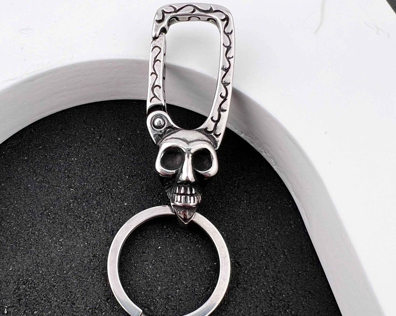 Puede incluir: Un llavero plateado con un colgante de calavera. La calavera tiene rasgos faciales detallados y est&aacute; unida a un llavero. La parte superior del llavero tiene un cierre decorativo con un dise&ntilde;o ornamental.