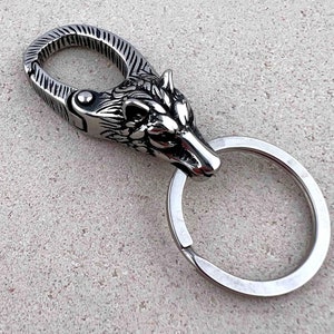 May include: A silver-colored keychain featuring a wolf head design. The keychain has a spring-loaded clasp and a split ring for attaching keys. The wolf head is intricately detailed, with a textured finish on the clasp.