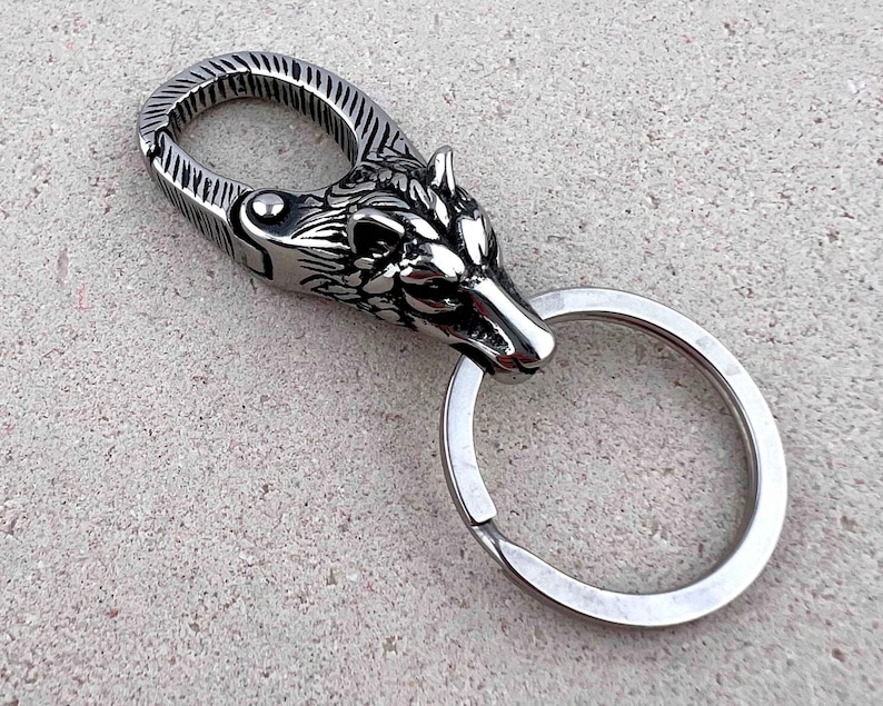 May include: A silver-colored keychain featuring a wolf head design. The keychain has a spring-loaded clasp and a split ring for attaching keys. The wolf head is intricately detailed, with a textured finish on the clasp.