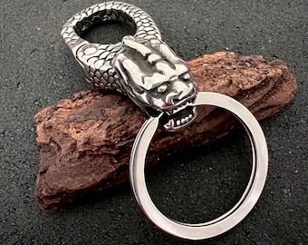 May include: A silver-colored keychain featuring a detailed dragon head design. The dragon's head is sculpted with scales and a fierce expression, holding a large, circular key ring. The keychain is made of a shiny, metallic material.
