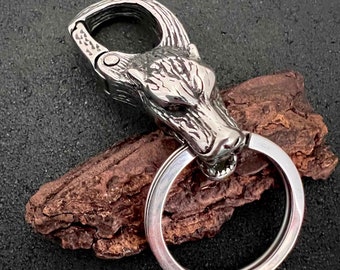 May include: A silver-colored keychain featuring a detailed wolf head design. The keychain includes a sturdy clasp and a key ring. The wolf head is intricately carved, showcasing the animal's features. The overall design is metallic and rugged.