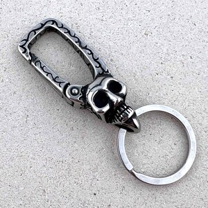 Puede incluir: Un llavero plateado con dise&ntilde;o de calavera. La calavera presenta cuencas de ojos huecas y una mand&iacute;bula prominente. El llavero tiene un cierre decorativo con un patr&oacute;n grabado y un llavero circular.