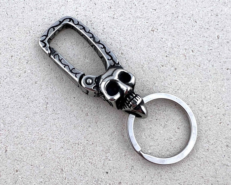 Puede incluir: Un llavero plateado con dise&ntilde;o de calavera. La calavera presenta cuencas de ojos huecas y una mand&iacute;bula prominente. El llavero tiene un cierre decorativo con un patr&oacute;n grabado y un llavero circular.