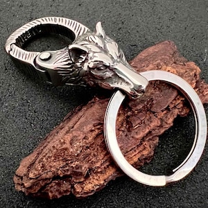 May include: A silver-colored keychain featuring a detailed wolf head design. The keychain includes a sturdy clasp and a large key ring. The wolf head is intricately sculpted, showcasing the animal's features. The overall design is metallic and rugged.