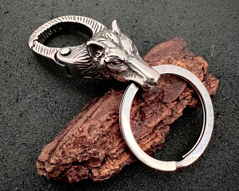 May include: A silver-colored keychain featuring a detailed wolf head design. The keychain includes a sturdy clasp and a large key ring. The wolf head is intricately sculpted, showcasing the animal's features. The overall design is metallic and rugged.